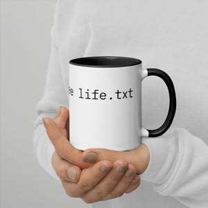 K&ouml;nnte beinhalten: Wei&szlig;e Keramik-Tasse mit schwarzem Innenraum, Henkel und dem Text "ee life.txt" in Schwarz. Die Tasse wird von einer Person gehalten, die einen wei&szlig;en Pullover tr&auml;gt.