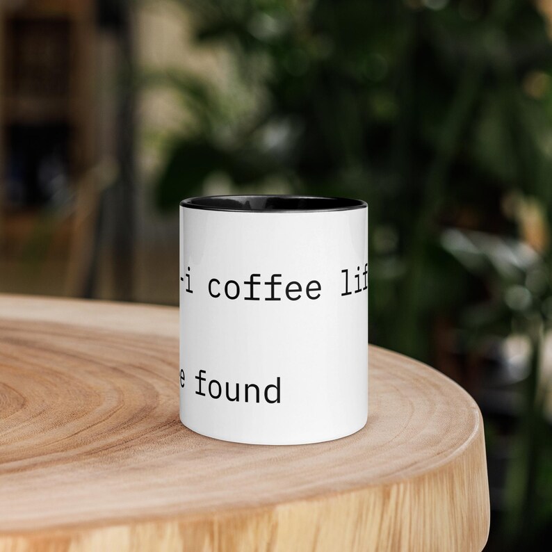 K&ouml;nnte beinhalten: Wei&szlig;er Kaffeebecher mit schwarzem Innenraum und dem Text "i coffee life" und "e found" in Schwarz gedruckt. Der Becher steht auf einer Holzoberfl&auml;che.