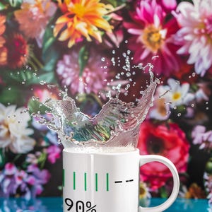 May include: A white ceramic mug with a green battery-like indicator showing 90% capacity. Water is splashing out of the mug. The background features a vibrant floral arrangement with pink, red, and yellow flowers.