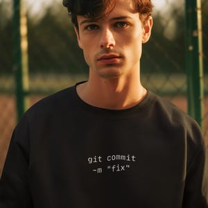 May include: A black sweatshirt with the text "git commit -m "fix"" in white. The person in the image has brown hair and is looking at the camera. The background is blurred.