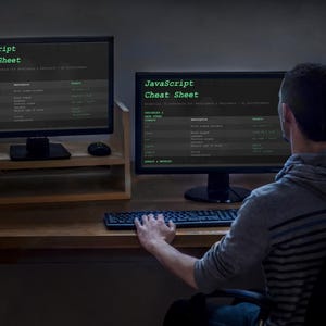 以下が含まれることがあります： 木製の机の上に「JavaScript Cheat Sheet」が表示された2台のコンピューターモニターがあります。キーボードを叩いている人がモニターの前に座っています。画面のテキストは緑色です。