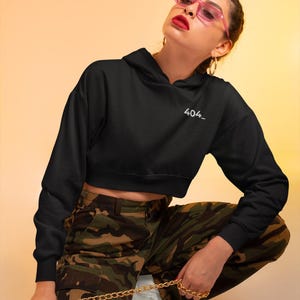 May include: Black cropped hoodie with the text "404_" in white. The model is wearing camouflage trousers and pink sunglasses. A gold chain is held in the hand. The background is a gradient of yellow and orange.