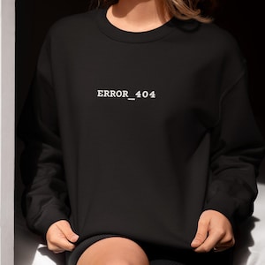 May include: Black crew neck sweatshirt with the text "ERROR_404" in white. The sweatshirt has long sleeves and a relaxed fit. The model is wearing black shorts. The overall aesthetic is casual and modern.