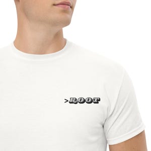 May include: White t-shirt with the word ">ROOT" printed in a 3D-style font on the front. The text is in a dark gray color with a black outline. The shirt is made of a soft material.