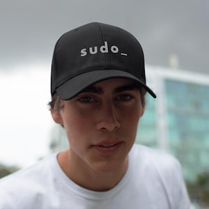 May include: A black baseball cap featuring the word sudo_ printed in white letters on the front panel. The cap has a structured crown, a curved brim, and a classic six-panel design with a button on top, providing a clean and simple look.