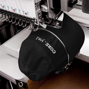 May include: A black baseball cap secured in an industrial embroidery machine. The cap features white embroidered text that reads CORE • ym1. The machine has multiple needles and thread guides positioned directly above the fabric of the cap.