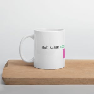 May include: White ceramic mug with the text "EAT. SLEEP. CODE" in black, green, and pink. The mug is on a wooden surface.