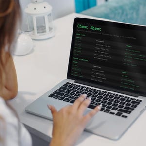 Pu&ograve; includere: Un laptop mostra una "Cheat Sheet" con testo verde su sfondo nero. Lo schermo mostra un elenco di comandi e descrizioni. Una mano &egrave; sulla tastiera. Il laptop &egrave; su una superficie bianca.