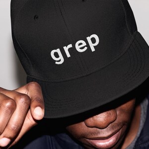 May include: Black baseball cap with the word "grep" embroidered in white. The cap has a curved peak and a structured fit. The person in the image is wearing a dark jacket.