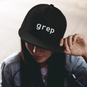 May include: Black flat-brimmed baseball cap with the word "grep" in white lettering. The cap is worn by a person, with the brim casting a shadow over their face. The person is wearing a denim jacket.