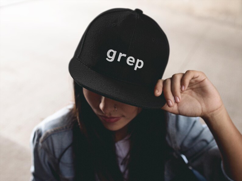 May include: Black flat-brimmed baseball cap with the word "grep" in white lettering. The cap is worn by a person, with the brim casting a shadow over their face. The person is wearing a denim jacket.