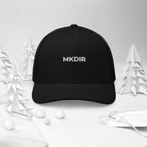 May include: Black trucker hat with the white text "MKDIR" embroidered on the front. The hat has a mesh back and a curved brim. The background features white paper Christmas trees and small white spheres.