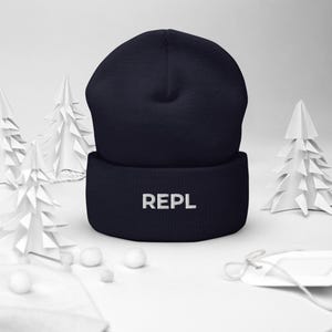 May include: A dark navy blue beanie with the word "REPL" embroidered in white on the folded brim. The hat is displayed on a white surface with decorative paper trees and small white spheres.