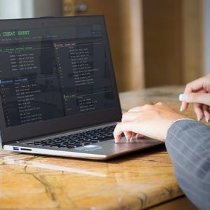 Pu&ograve; includere: Un laptop mostra una "VIM CHEAT SHEET" sullo schermo. Lo schermo mostra righe di codice e comandi in testo verde e bianco. Il laptop &egrave; aperto su un tavolo di legno, con le mani di una persona che digita sulla tastiera.