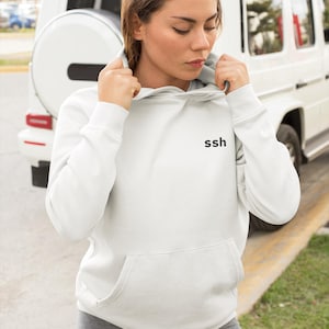 May include: A white hoodie with a front pocket and the letters "ssh" printed on the chest. The model is wearing gray leggings. A white SUV is in the background.