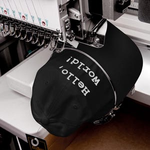 May include: A black baseball cap mounted on an industrial embroidery machine, featuring the white embroidered text "Hello, World!" on the front panel. The machine includes multiple needles and thread spools positioned above the cap.