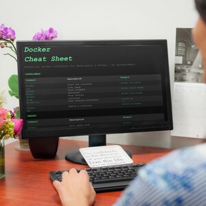 May include: A computer monitor shows a "Docker Cheat Sheet" with commands and descriptions. A keyboard and a small card with text are in front of the monitor. A vase of flowers is on the left. The scene is set on a wooden desk.