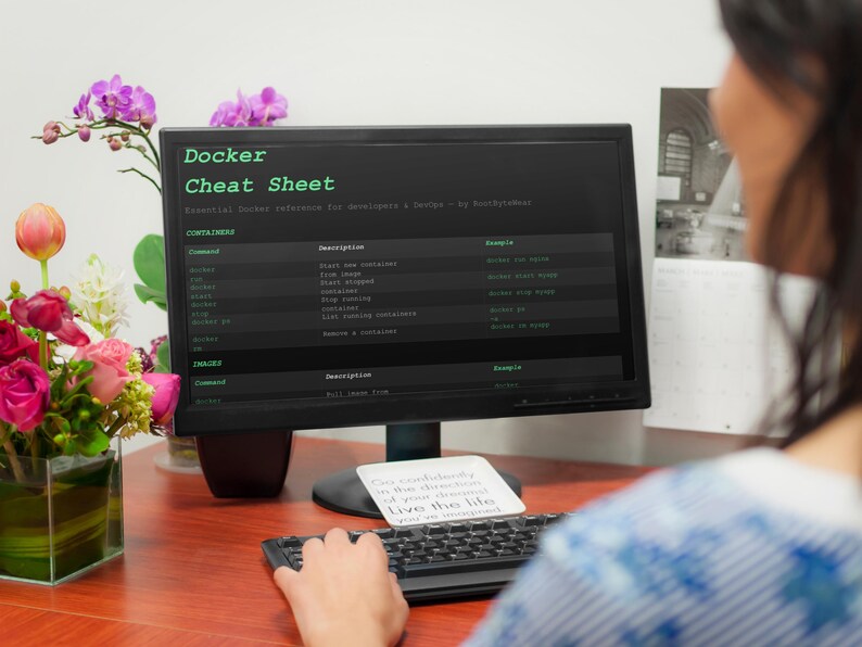 May include: A computer monitor shows a "Docker Cheat Sheet" with commands and descriptions. A keyboard and a small card with text are in front of the monitor. A vase of flowers is on the left. The scene is set on a wooden desk.