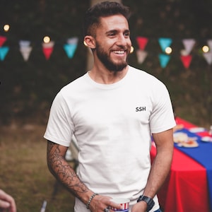 May include: A white short-sleeved t-shirt featuring the letters SSH printed in black on the left side of the chest. The garment has a crew neckline and a standard fit, constructed from a plain white fabric with no additional patterns or designs.