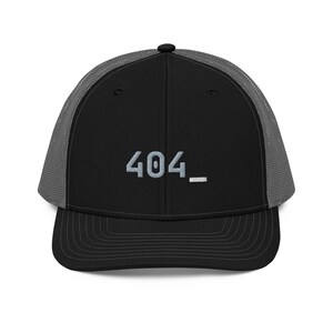 May include: Black trucker hat with a gray mesh back and a curved brim. The front panel displays the embroidered text "404_" in a light gray color. The hat has a structured fit and a snapback closure.
