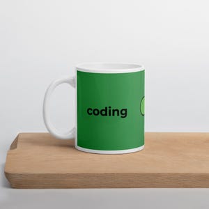 May include: White ceramic mug with a green background and the word "coding" in black. A green circle is next to the word. The mug is sitting on a wooden surface.