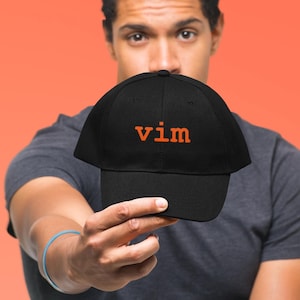 May include: A black baseball cap featuring the word vim embroidered in orange lowercase letters on the front panel. The cap has a structured crown, a curved brim, and a button at the top center. The fabric appears to be a solid black cotton.