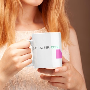 May include: White ceramic mug with the text "EAT. SLEEP. CODE." in black and green. A pink square is next to the word "CODE." The mug is held by a person with long, light-colored hair.