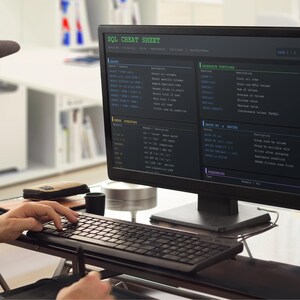 以下が含まれることがあります： 帽子とメガネをかけた人がパソコンで作業しています。モニターにはコードスニペット付きの「SQL Cheat Sheet」が表示されています。キーボード、マウス、その他のアクセサリーが机の上にあります。