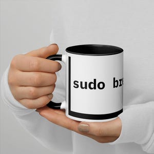 May include: White ceramic mug with a black interior and handle. The mug features the words "sudo br" in black, sans-serif font. The mug is held by a person wearing a white long-sleeve shirt.