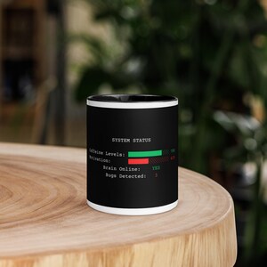 May include: A black ceramic mug with a white interior and rim. The exterior features white text reading SYSTEM STATUS, Caffeine Levels, Motivation, Brain Online: YES, and Bugs Detected: 3, alongside a green bar for caffeine and a red bar for motivation.