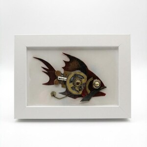 Op de afbeelding: Een steampunk-stijl vissculptuur in een witte lijst. De vis is gemaakt van metalen tandwielen, platen en klinknagels, met een kleurenpalet van brons, koper en bordeauxrood. Het kunstwerk staat tegen een witte achtergrond.