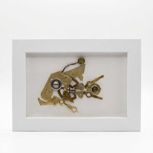 May include: A framed artwork depicting a fish made from brass and gold-toned metal components. The fish is set within a white frame, offering an industrial aesthetic. The artwork is a creative blend of mechanical parts, presented in a clean, modern style.