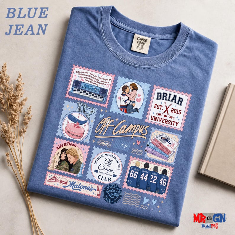 a blue t-shirt with various colorful and whimsical designs, including a couple embracing, a piano, and other decorative elements. The t-shirt is laid out on a surface, and there are some decorative elements like a book and a plant nearby.