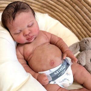 May include: A lifelike baby doll with closed eyes, resting on a white surface. The doll has a realistic skin tone, dark hair, and is wearing a diaper with the text "Millie Moon". A stuffed bunny is in the background.