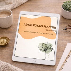 Może przedstawiać: Biały tablet wyświetla okładkę ADHD Focus Planner 2026 Edition. Okładka zawiera pomarańczowy abstrakcyjny kształt, rysunek kwiatu i tekst "Minimalistyczny system dla jasności i produktywności."