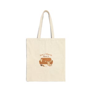 May include: Cream-colored tote bag with a cartoon illustration of a brown couch, blanket, and movie accessories. The text "Home is Where the Movie Is" is printed above the couch. The bag has two cream-colored handles.