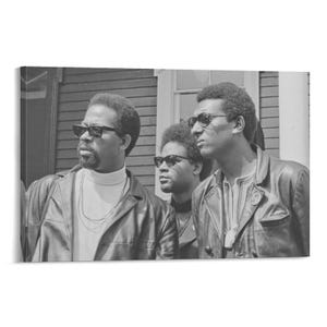 May include: Black and white photograph of three men wearing sunglasses and leather jackets. The men are standing outside a building. One man is wearing a turtleneck and a chain necklace.