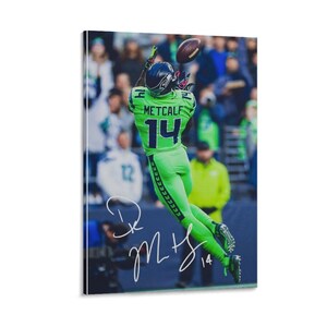 Op de afbeelding: Een ingelijste print met een American footballspeler in een groen uniform met nummer 14, die een bal vangt. De naam van de speler, Metcalf, is zichtbaar. De print is gesigneerd. De achtergrond toont een menigte.