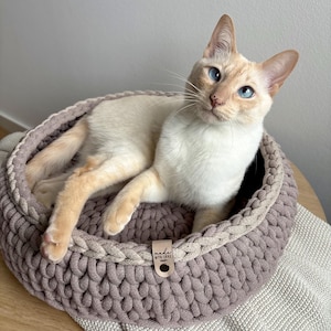 Op de afbeelding: Een crèmekleurige kat met blauwe ogen rust in een rond, geweven kattenbed. Het bed is een gedempt paars met een lichtere beige rand. Een klein label op het bed luidt "made with love". De kat ligt op een witte deken.