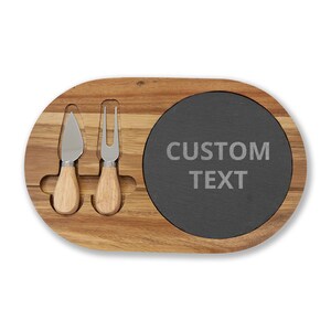 Könnte beinhalten: Käsebrett aus Holz mit einer Schiefer-Servierfläche und zwei Käsewerkzeugen. Das Brett hat eine natürliche Holzmaserung. Der Schieferbereich trägt die Aufschrift "CUSTOM TEXT".