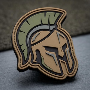 May include: A tan, green, and black patch featuring a stylized Spartan helmet design. The helmet has a detailed crest and face guard, with a black border. The patch is likely made of durable material, suitable for attachment to clothing or gear.
