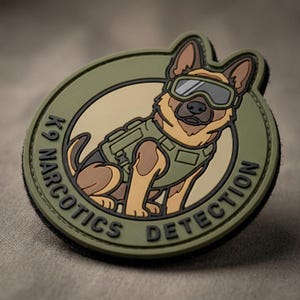 May include: A circular olive green tactical patch featuring a cartoon German Shepherd dog wearing a tactical vest and goggles. The text K9 NARCOTICS DETECTION is embossed in black along the bottom edge of the patch, which has a raised rubberized texture.