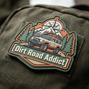 May include: A rectangular PVC patch with rounded edges featuring a graphic of an off-road vehicle, pine trees, mountains, and a compass rose. The text Dirt Road Addict is printed in white on an orange background. The patch has green borders.