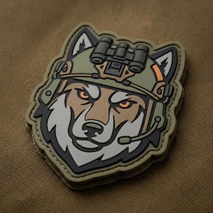 May include: A patch featuring a wolf's head wearing a military helmet with night vision goggles. The patch has a tan, green, and black color scheme with orange eyes. The wolf has a serious expression and a headset.