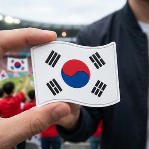May include: A white patch featuring the South Korean flag, held in front of a blurred background of a crowd. The flag has a central yin-yang symbol in red and blue, surrounded by four black trigrams.