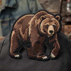 May include: An embroidered patch featuring a brown grizzly bear in a walking pose. The bear has detailed fur textures in shades of dark brown, tan, and cream, with a black stitched border outlining the entire shape against a dark fabric background.