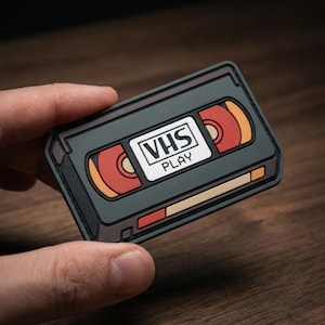 May include: A rectangular PVC patch shaped like a vintage VHS cassette tape. The design features a dark gray frame with red and orange circular reel details. A central white label displays the black text VHS and PLAY in a bold, sans-serif font.