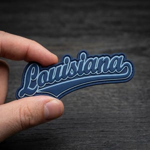May include: A blue and white Louisiana state souvenir patch held in a hand. The patch features the word "Louisiana" in a stylized script font with a white outline and a blue background. The patch is a souvenir item.