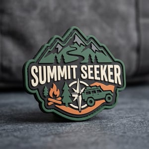 May include: A mountain-shaped morale patch featuring the text SUMMIT SEEKER in cream lettering. The design includes a green mountain range, a campfire, pine trees, a compass rose, and an off-road vehicle in shades of green, orange, and black.