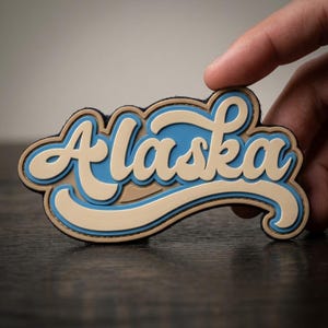 May include: A layered Alaska souvenir patch in a retro style. The word "Alaska" is in a cursive font, with a light blue and tan color scheme. The patch is held by a person's fingers.